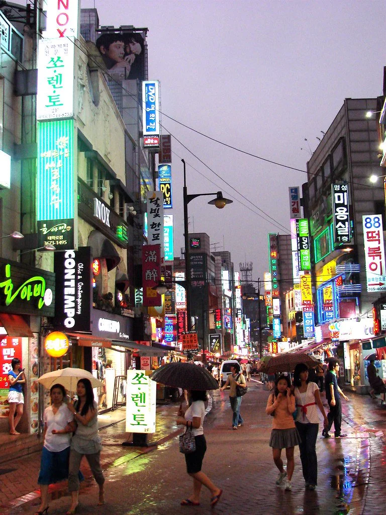 비 내리는 밤 서울 도심 거리에서 우산을 쓴 사람들이 네온 간판 아래를 걷고 있다
